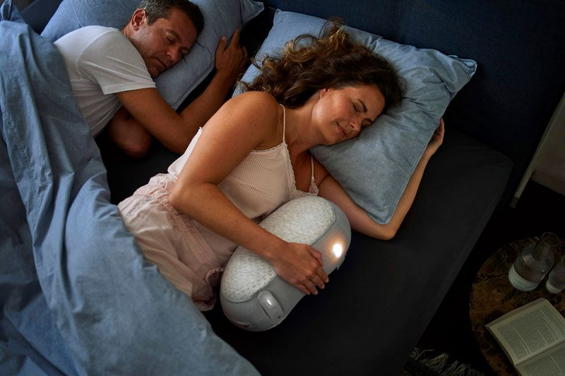 Fotografie eines schlafenden Paares. Frau hält den Somnox-Roboter.